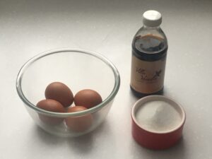 A glass bowl with 4 eggs, a dish with sugar, and a bottle of vanilla